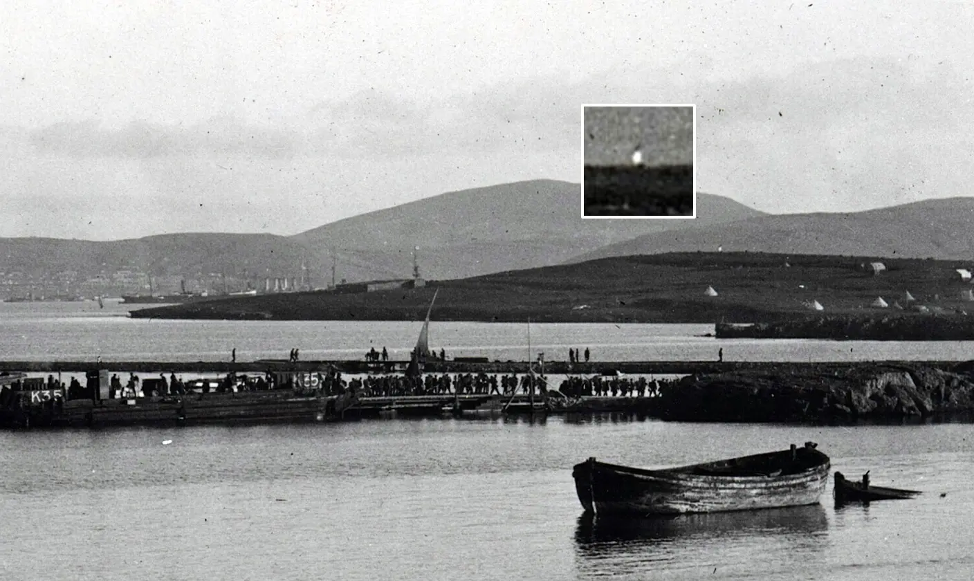 A black and white photo shows a calm lake with two wooden boats in the foreground. In the background, hills under a cloudy sky. A small bright spot is highlighted.
