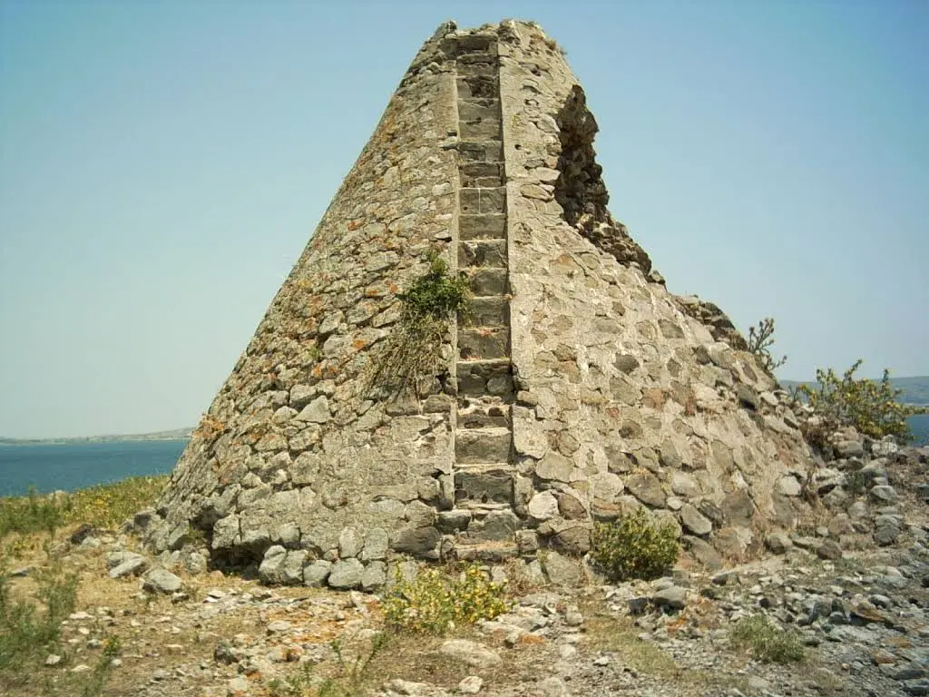 Stone pyramid-shaped structure with a central vertical indentation and scattered plants, set on a rocky hillside near a calm blue sea under a clear sky.