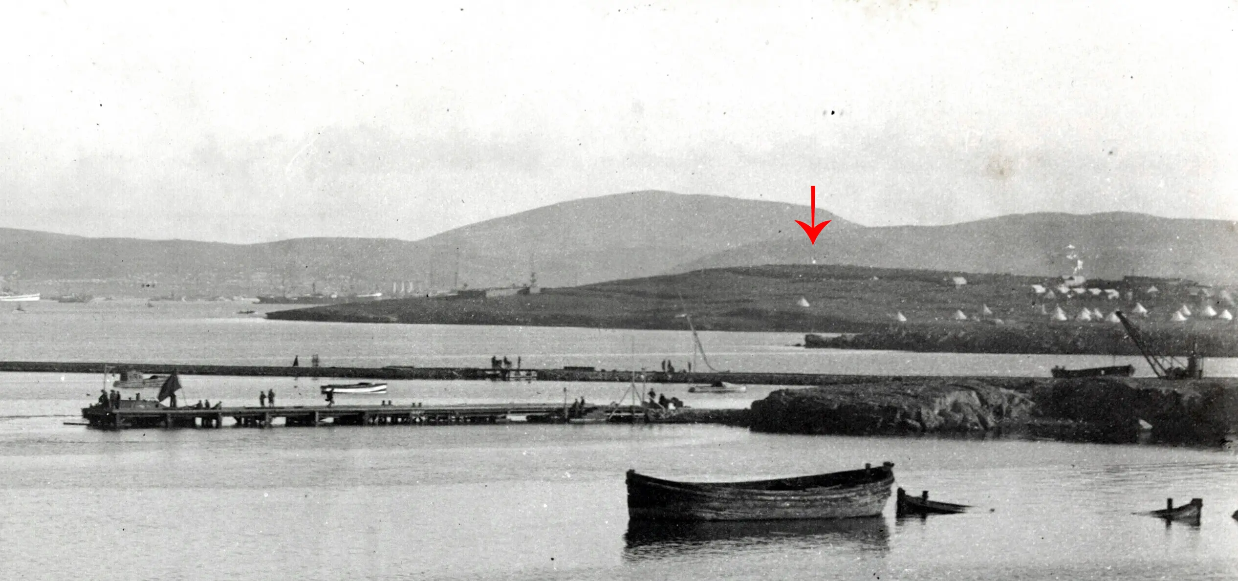 Calm waterfront scene with several small boats floating. A distant hill with a row of white tents sits under a cloudy sky, creating a serene atmosphere. A small bright spot is highlighted on one of the distant hills.