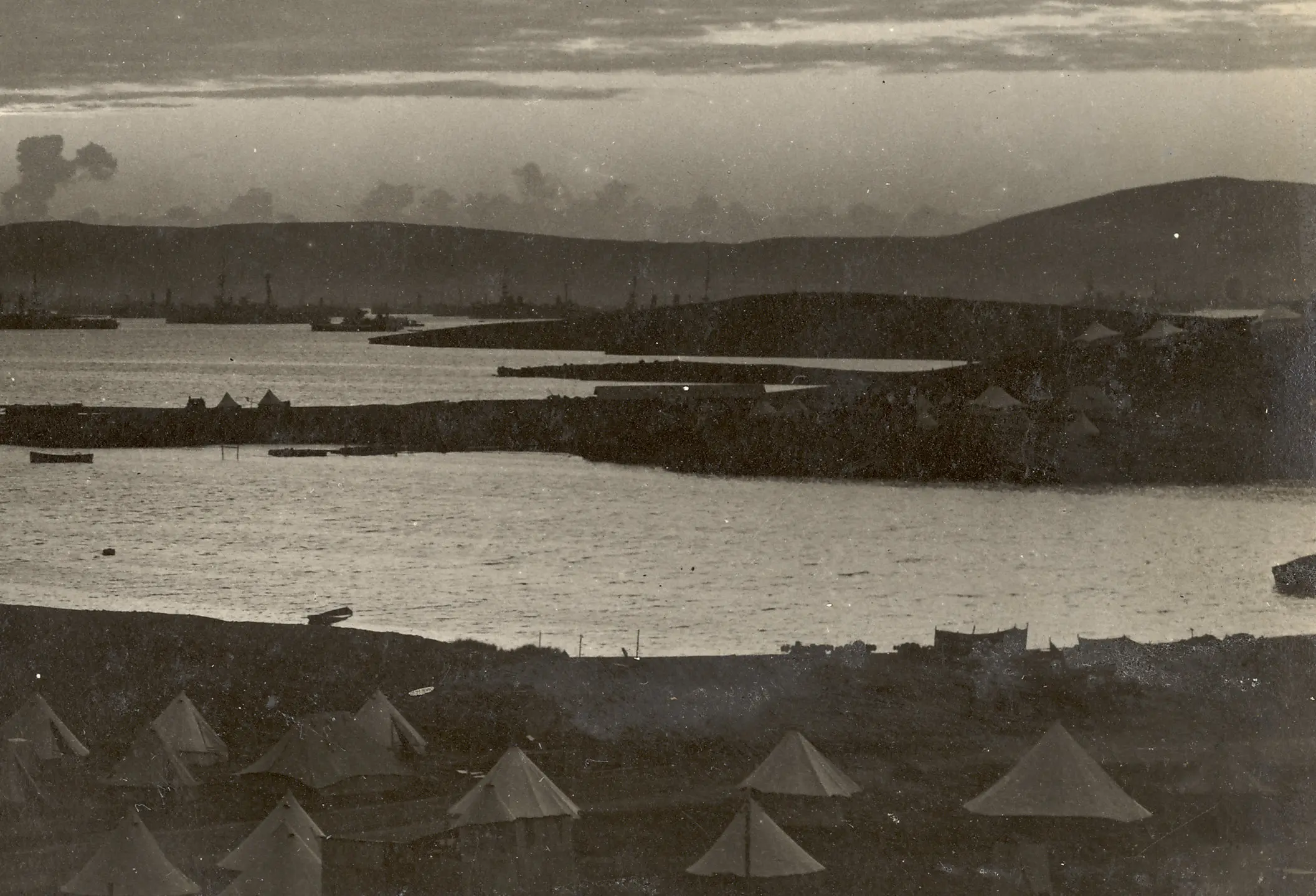 A serene, sepia-toned scene of a bayside camp at dusk. Canvas tents cluster in the foreground, while calm waters and a hilly skyline fill the distance.