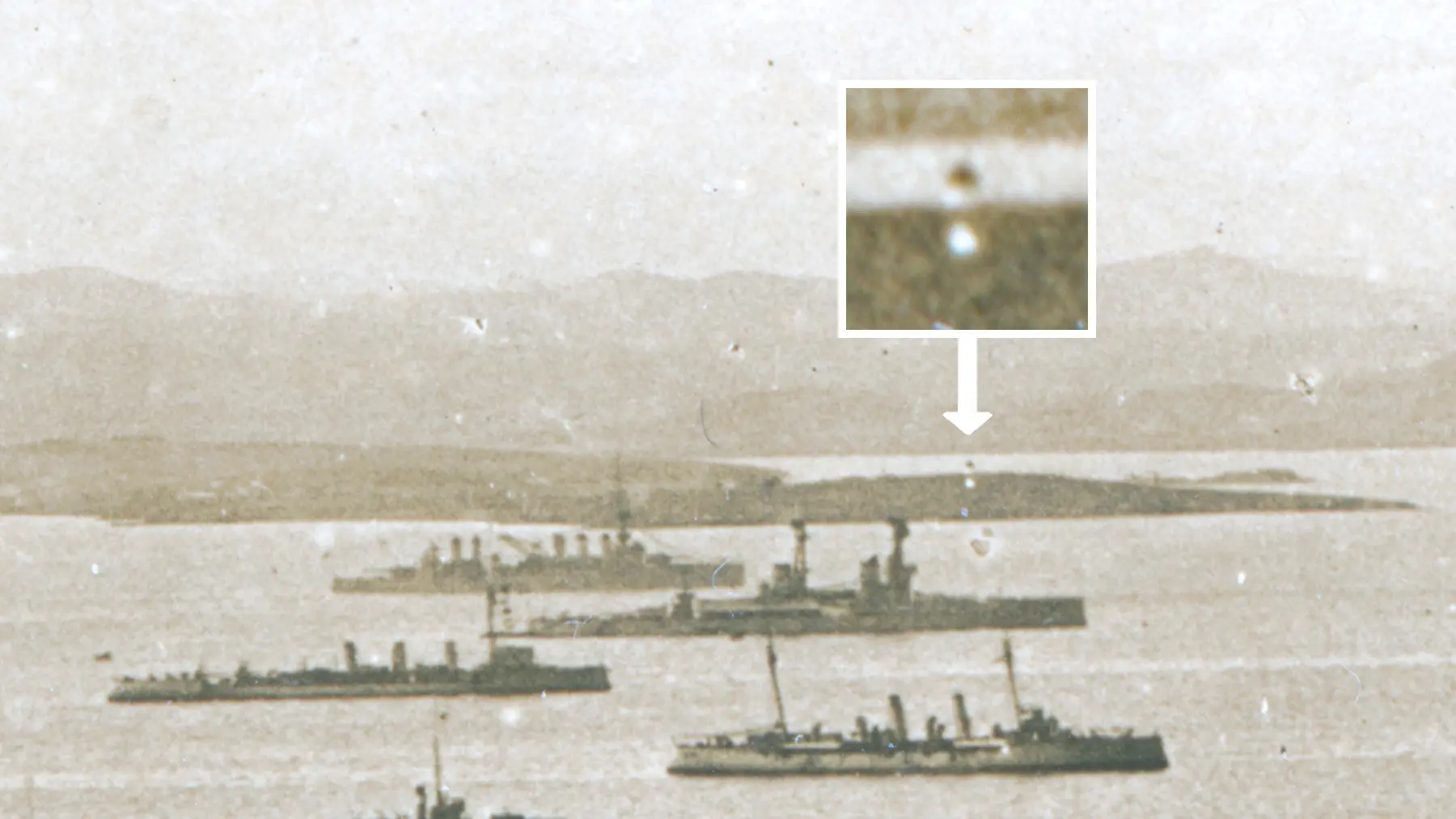 Sepia-toned image of several ships in a harbor with distant hills. Inset shows a magnified view of a distant object in the water, highlighted for focus.
