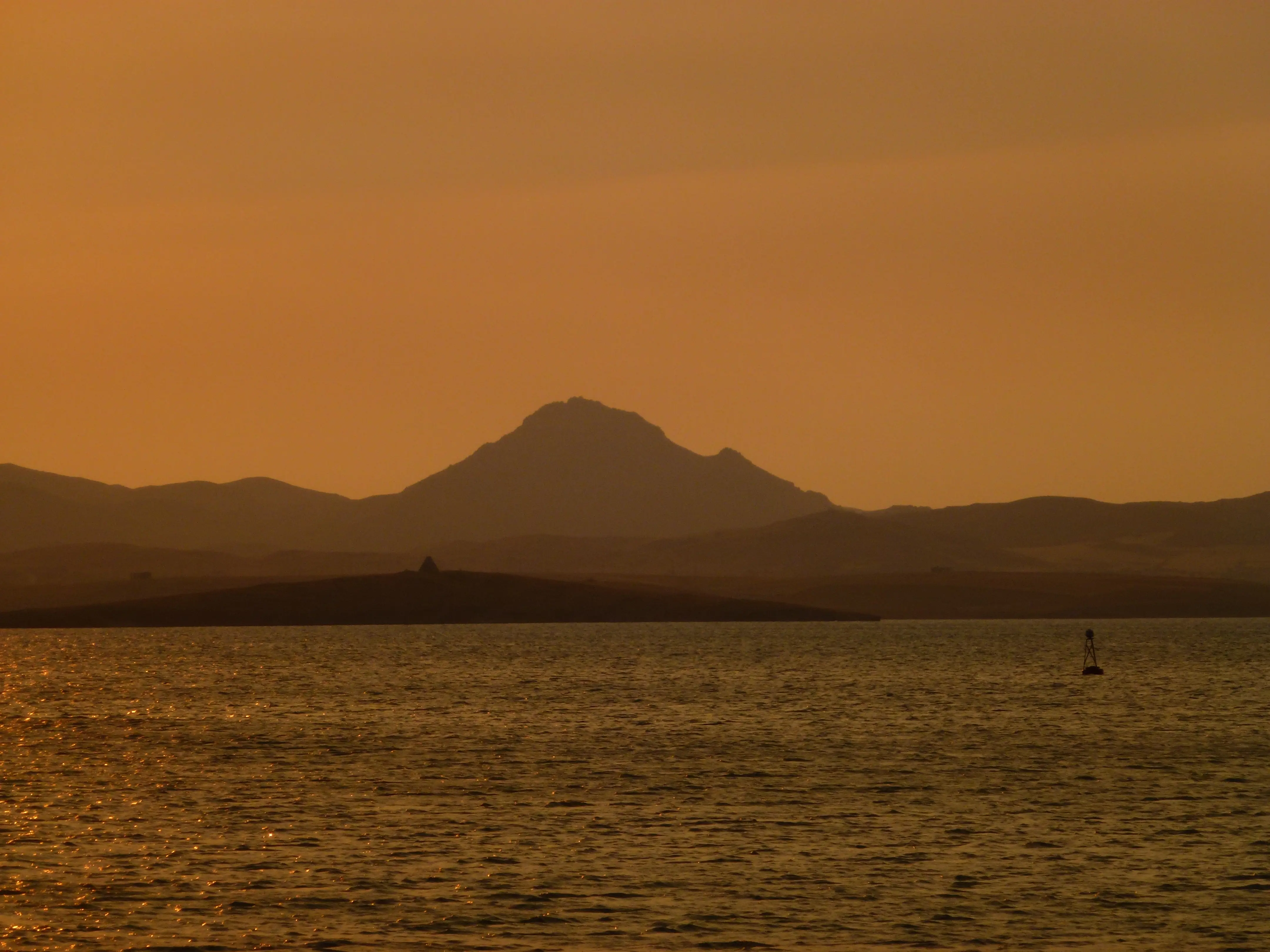 Sunset over tranquil water with gentle ripples. A dark silhouetted mountain range in the background under a warm golden sky. In the foreground, low land with a pyramid-like structure at its summit.