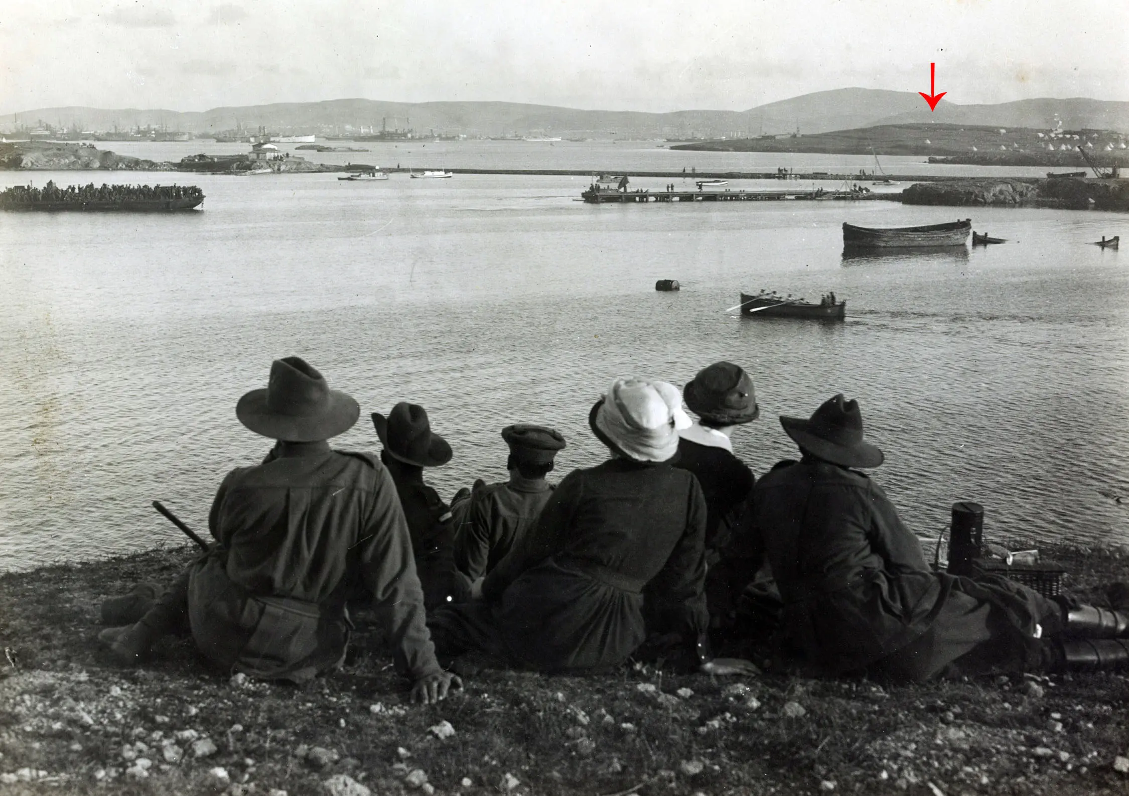 A group of people sit on a grassy hill, watching ships and boats on a calm body of water. Hills are visible in the distance under a cloudy sky. A small bright spot is highlighted on one of the distant hills.