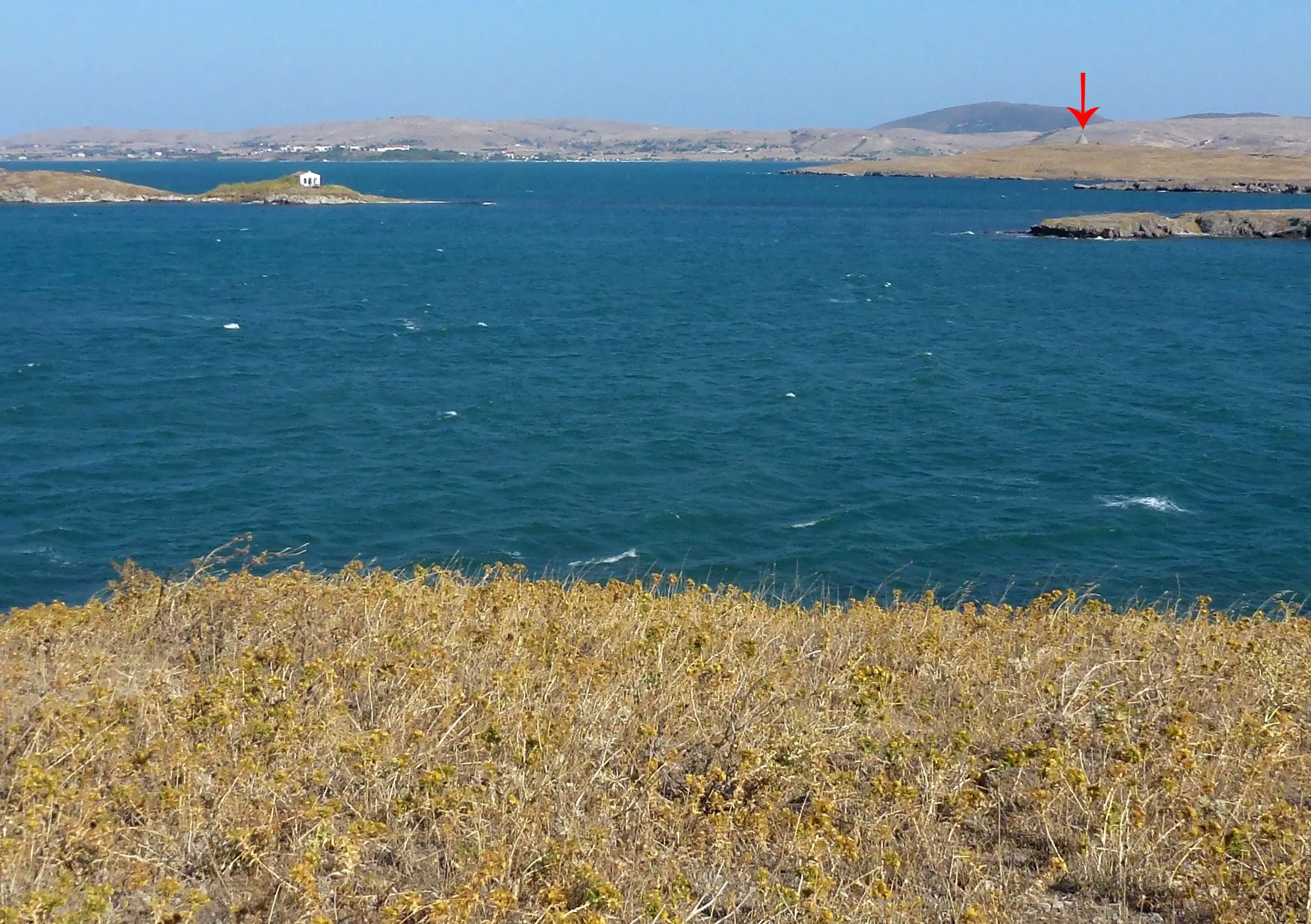 Coastal landscape with a foreground of dry grass, a deep blue sea, and distant hills under a clear sky. A small chapel is visible on a distant island. A small bright spot is highlighted on one of the distant hills.