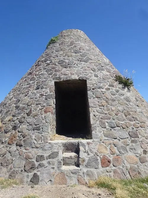 A stone structure shaped like a cone against a clear blue sky, with a dark rectangular doorway and small plants growing on its side.